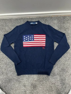 Mörkblå stickad tröja Polo Ralph Lauren - Mörkblå stickad tröja från Polo Ralph Lauren med amerikansk flagga i rött, vitt och blått på bröstet. RL-broderi i rött nedanför flaggan. Tröjan har rund halsringning, långa ärmar och ribbade muddar. Perfekt för dig som gillar klassisk college-stil.