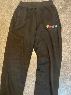 Svarta mjukisbyxor Trapstar - Svarta mjukisbyxor från Trapstar med broderad logga i flera färger på vänster lår. Byxorna har resår i midjan och sidofickor. Perfekta för en avslappnad och trendig stil.