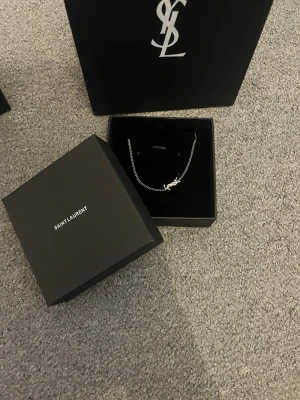 Silver armband från Yves Saint Laurent - Snyggt silverfärgat armband från Saint Laurent med den ikoniska YSL-loggan som hänge. Kedjan är tunn och elegant, perfekt för att lyfta din outfit med en lyxig touch. Kommer i originalask med Saint Laurent-logga.