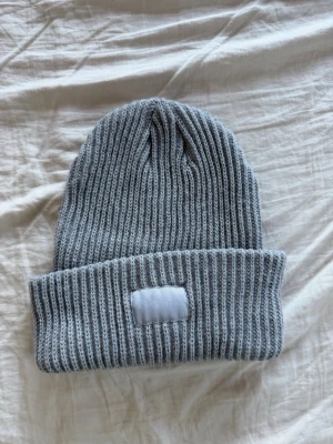 Ljusgrå ribbstickad mössa - Säljer en stilren ljusgrå ribbstickad mössa med uppvikt kant och en vit patch framtill. Mössan har en klassisk form och är tillverkad i mjukt, stretchigt material som håller dig varm. Perfekt accessoar för kyliga dagar.