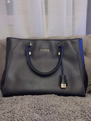 Svart handväska från Guess - Snygg svart handväska från Guess med guldfärgade detaljer och logotyp framtill. Använd endast 3-4 gånger köptes för 2199