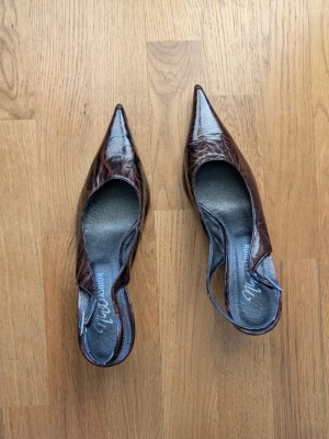 Bruna spetsiga pumps i lack - Säljer ett par bruna pumps från Marco Tozzi med spetsig tå och smal klack. Skorna har en glansig lackyta och öppen häl med rem runt ankeln. Perfekta för dig som gillar en elegant och trendig look.