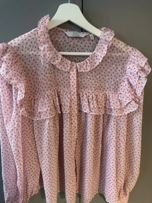 Ljusrosa volangblus med prickar - Söt ljusrosa blus från & Other Stories med små prickar i metallic och volangdetaljer vid axlar och bröst. Blusen har rundad krage, knäppning framtill och är gjord i ett tunt och transparent tyg. 