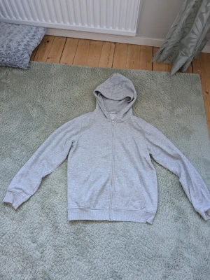 Ljusgrå zip-hoodie med huva - En stilren ljusgrå hoodie med dragkedja framtill och huva. Tröjan har långa ärmar, ribbade muddar och två fickor framtill. Perfekt för en avslappnad look. 