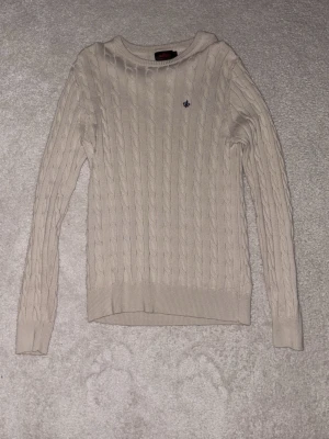 Beige kabelstickad tröja Morris - Kabelstickad tröja i beige från morris med rund halsringning och diskret logga broderad på bröstet. Tröjan har långa ärmar och ribbade muddar vid ärmslut och nederkant. Perfekt för lager på lager och klassisk stil.