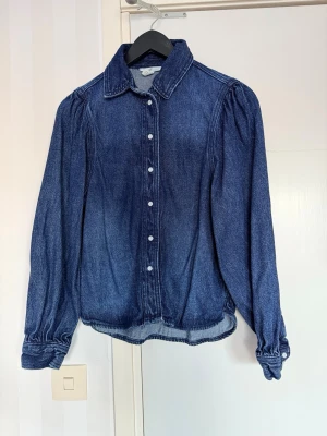Mörkblå jeansskjorta från H&M - Snygg mörkblå jeansskjorta från H&M i 100% bomull. Skjortan har puffiga axlar, klassisk krage och knappar framtill. Ärmsluten har knappar och skjortan har en avslappnad passform som ger en cool vintagekänsla.