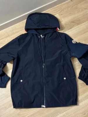 Svart vindjacka från Moncler - Snygg mörkblå vindjacka från Moncler med huva och dragkedja framtill. Jackan har två fickor med tryckknappar och Moncler-logga på ärmen. ,Perfekt för blåsiga dagar. Priset är ej hugget i sten!