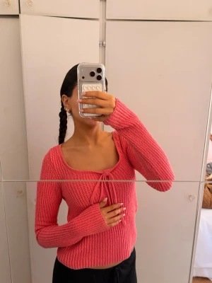 Rosa stickad tröja med knytning - Trendig rosa stickad tröja med lång ärm och snygg knytning som man själv kan knyta framtill vid den breda, fyrkantiga urringningen. Tröjan är ribbstickad och har en figurnära passform som ger en modern look. Perfekt för dig som gillar färg och vill sticka ut. 