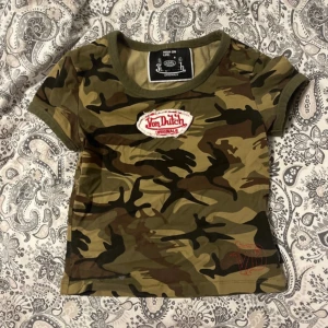 Camo crop topp från Von Dutch - Cool camouflagemönstrad topp från Von Dutch med korta ärmar och rund halsringning. T-shirten har en tydlig logopatch framtill och är i en tight passform. Perfekt för dig som vill sticka ut med en streetstyle-look.