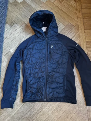 Svart hybriddunjacka från Peak Performance - Snygg svart dunjacka från Peak Performance med huva och hel dragkedja. Jackan har quiltad front, bröstficka med dragkedja och diskret logga på bröstet. 