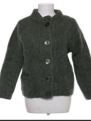 Mörkgrön stickad kofta med stora knappar - Mysig mörkgrön stickad kofta med stora knappar framtill och rak passform. Koftan har lång ärm och en hög krage som ger extra värme. Perfekt för lager på lager och en chill look. Jag har tagit bort lappen så jag minns tyvärr inte var den är i från men materialet är väldigt fint. 