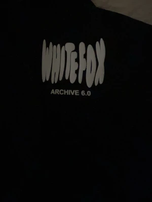 Svart hoodie från White Fox - Snygg svart hoodie från White Fox med trycket 'WHITE FOX ARCHIVE 6.0' i vitt framtill. Oversized passform och klassisk huva. Perfekt för dig som gillar streetwear och vill ha en enkel men trendig look. Köpt utomlands!