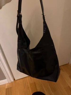 Svart axelväska i fejkläder - Stilren svart axelväska i mjukt fejkläder med enkel design och bred axelrem. Väskan har en rymlig insida och passar perfekt för dig som gillar minimalistisk stil. Perfekt att slänga över axeln till skolan eller stan.