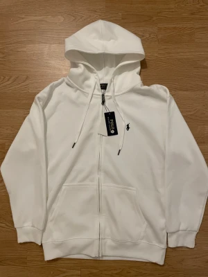 Vit hoodie från Polo Ralph Lauren - Snygg vit hoodie med dragkedja från Polo Ralph Lauren. Klassisk design med huva, dragsnören och känguruficka framtill. Liten broderad logga på bröstet. Perfekt för en clean och stilren look. Passar M 