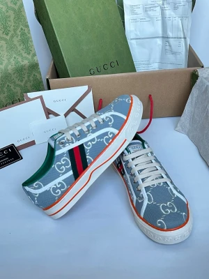 Gucci GG Supreme sneakers grå - Snygga sneakers från Gucci i grått tyg med vitt GG Supreme-mönster, gröna och röda sidoremsor samt orange detalj på sulan. Klassisk låg modell med vita snören och grönt innerfoder. Perfekta för dig som vill sticka ut med exklusiv streetstyle.