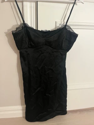 Svart kort klänning från Zara - Snygg svart kort klänning från Zara med tunna axelband och spetsdetaljer vid bysten. Överdelen har diskreta ränder och kjolen är slät. Perfekt för dig som gillar en enkel men elegant look.