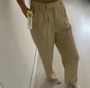 Beige kostymbyxor med snygg midjs  - Ett par helt oanvända beige kostymbyxor (prislappkvar) Dessa är endast testade för att ta bilden till försäljning. Knäpps med dragkedja, knapp och en extra hake-vilket gör midjan är ursnygg! (Liknar madlady’s kostymbyxor) 
