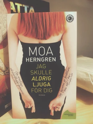 Jag skulle aldrig ljuga för dig 🤞 - Romanen 'Jag skulle aldrig ljuga för dig' av Moa Herngren är en pocketbok med ett omslag som visar en person med rött hår i svart klänning och text på armarna. Boken är tryckt på papper och har en modern, ungdomlig design.