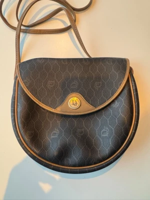 Christian Dior axelväska – vintage - Elegant Christian Dior-väska, tillverkad i Frankrike. Klassisk grå/beige monogramdesign med beige kantdetaljer och justerbar axelrem. En tidlös modell som passar både vardag och mer uppklädda tillfällen.  Väskan är i begagnat skick med tydliga bruksspår (se bilder): viss slitage och missfärgning i materialet samt patina på detaljerna, vilket är vanligt för vintage Dior och ger väskan karaktär. Stängning med knapp, lätt och smidig storlek – perfekt som crossbody.  Detaljer: 	•	Märke: Christian Di