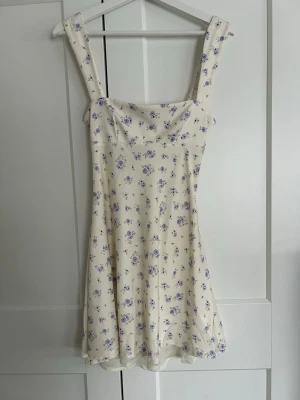 Blommig kort klänning med rosett - Söt kort klänning i ljus cremevit färg med lila blommönster. Klänningen har breda axelband och en fin rosettdetalj i ryggen. Perfekt för varma dagar och har en feminin, lätt utsvängd passform. Oanvänd med lappen kvar🌸