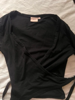 Svart omlottopp från Nelly - Svart omlottopp från Nelly med v-ringning och knyt i sidan. Toppen har korta ärmar och är tillverkad i mjuk bomull, perfekt för en trendig och avslappnad look.