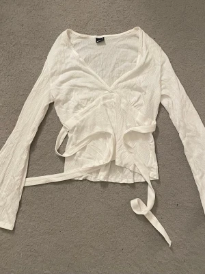 Vit topp från Gina Tricot storlek M - Säljer en vit topp från Gina Tricot med långa ärmar och knytband i midjan. Toppen har v-ringning och är gjord i ett lätt, krinklat material som känns luftigt och skönt. Perfekt att styla med jeans eller kjol.