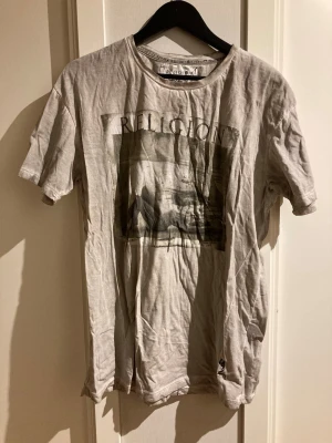 Grå t-shirt från Religion med tryck - Säljer en grå t-shirt från Religion med ett stort grafiskt tryck på bröstet och texten 'RELIGION' ovanför motivet. T-shirten har rund halsringning och korta ärmar. Perfekt för dig som gillar streetwear och vill ha en unik look.
