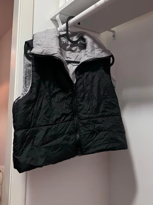 Svart dunväst med grå insida - Snygg svart dunväst med kontrasterande ljusgrå insida och hög krage. Västen har dragkedja framtill och quiltad design som ger en sportig look. Perfekt att slänga över en hoodie eller tröja för extra värme. Materialet är glansigt och lätt. Sitter liter nedanför midjan 