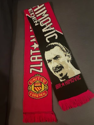 Zlatan Ibrahimovic supporterscarf - Röd och svart halsduk med texten 'Zlatan Ibrahimovic' Med Zlatans ansikte på och Manchester Uniteds logga på. 