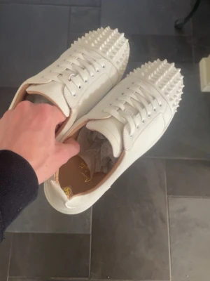Vita Christian Louboutin sneakers med nitar - Snygga vita sneakers från Christian Louboutin med ikoniska röda sulor och coola nitar på tån och sidorna. Skorna är i läder och har vita skosnören. Perfekta för dig som vill sticka ut med en lyxig och edgy stil.