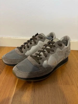 Grå sneakers från Philippe Model - Snygga sneakers i grått från Philippe Model med ovandel i mocka och mesh. Skorna har mörkgrå snörning, diskret logga på sidan och svart sula. Perfekta för en stilren och avslappnad look. 