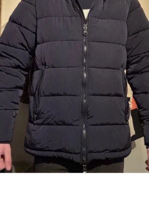 Mörkblå pufferjacka med dragkedja - Snygg mörkblå pufferjacka med quiltad design och två sidofickor med dragkedja. Jackan har hög krage och en rak passform som ger en modern look. Perfekt för kalla dagar när du vill hålla värmen och ändå se stilren ut.