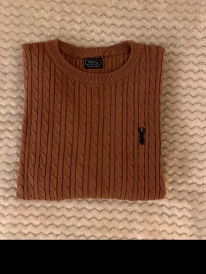 Brun kabelstickad tröja från Next - Säljer en brun kabelstickad tröja från Next med rund halsringning och diskret broderad logga på bröstet. Tröjan har långa ärmar och ribbade muddar vid ärmslut och nederkant. Perfekt för dig som gillar klassisk och stilren design.