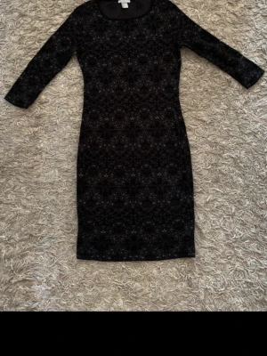 Svart mönstrad klänning med trekvartsärm - Snygg svart klänning med diskret mönster och trekvartsärm. Klänningen har en kroppsnära passform och rund halsringning. Perfekt för dig som gillar stilrena detaljer och enkel elegans.