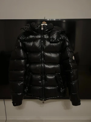 Svart moncler maya i stl 2 - Snygg svart dunjacka från Moncler med glansig finish och quiltad design. Jackan har hög krage, dragkedja framtill, fickor med dragkedja och Moncler-logga på ärmen. Perfekt för kalla dagar och ger en riktigt trendig look. Stl 2, en av de spännerna vid midjan har gått av, men de är inget man märker!