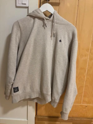 Beige morris hoodie i storlek M - Beige hoodie från morris med broderad liten logga på bröstet och patch nertill. Klassisk modell med huva och dragsko, ribbade muddar vid ärmslut och nederkant. Mjuk och skön bomullsblandning, perfekt för en avslappnad stil. Mycket bra skick då hoodien inte kommit till så mycket använding