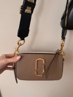 Marc Jacobs Snapshot axelväska beige - Säljer en beige Marc Jacobs Snapshot axelväska med guldfärgade detaljer och bred svart axelrem med logga. Väskan har en rektangulär form, dragkedja upptill och dekorativ tofs. Perfekt för dig som vill ha en trendig och stilren accessoar.