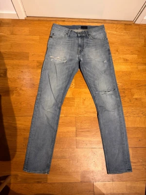 Ljusblå slitna jeans från Tiger of Sweden - Säljer ett par ljusblå jeans med raka ben och slitningar på både lår och knä. Klassisk femficksmodell med dragkedja och knapp. Jeansen har en avslappnad vibe och passar perfekt till en streetwear-look.