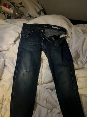 Replay Anbass mörkblå  - Snygga mörkblå Replay Anbass jeans med slitning på ena benet. Klassisk femficksmodell med knappgylf och smal passform. Perfekta för dig som gillar en modern och stilren look. Materialet är mjukt denim i bomull.