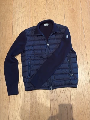 Mörkblå Cardigan från Moncler - Snygg mörkblå Cardigan från Moncler med stickade ärmar och quiltad front. Cardiganen har två dragkedjeförsedda fickor och klassisk Moncler-logga på ärmen. Perfekt för lager-på-lager och streetstyle. Materialmix av dun och bomull för både värme och stil. Den är i st 3(L)