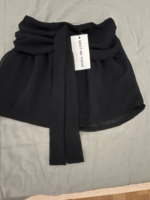 Svart crop top från Meet Me There XS/XXS - Trendig svart crop top från Meet Me There med omlottkänsla och bred knytband framtill. Tillverkad i ett lätt, flödande tyg som ger en snygg siluett. Perfekt att styla med höga jeans eller kjol för en cool look. Aldrig använd 