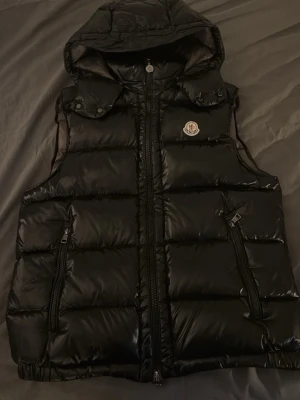 Svart dunväst från Moncler med huva - Snygg svart dunväst från Moncler med avtagbar huva och två dragkedjeförsedda fickor. Västen har en glansig finish, quiltad design och Moncler-logga på bröstet. Perfekt för lager-på-lager och streetwear-stil.