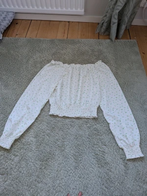 Vit offshoulder blus med blommönster - Söt vit offshoulder blus med små gröna blommor. Blusen har långa ärmar med resår vid ärmslut och midja, vilket ger en fin volangkant. Perfekt för en romantisk och trendig look. Tunn och luftig i materialet. Knappt använd.