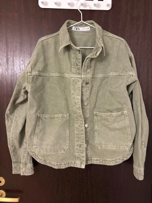 Olivgrön jeansjacka från Zara - Stilren olivgrön jeansjacka från Zara med klassisk krage, stora framfickor och knäppning framtill. Jackan har en avslappnad passform och raka ärmar, perfekt för en trendig vardagslook.