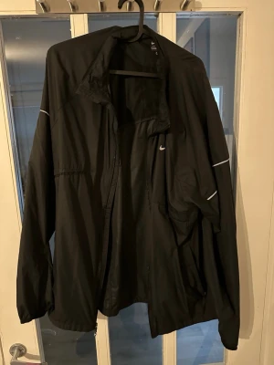 Svart vindjacka från Nike - Snygg svart vindjacka från Nike med dragkedja framtill och diskreta reflexdetaljer på ärmarna. Jackan har hög krage, elastiska muddar och en liten vit Nike-logga på bröstet. Tillverkad i lätt och tåligt material, perfekt för blåsiga dagar.