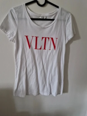 Vit VLTN t-shirt från Valentino - Säljer en vit t-shirt från Valentino med röd VLTN-logga framtill. Klassisk rund halsringning och korta ärmar. Tillverkad i 100% bomull för en skön och luftig känsla. Perfekt för dig som gillar stilrena och trendiga plagg.