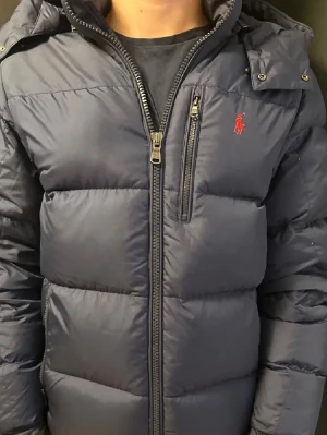 Mörkblå pufferjacka från Ralph Lauren - Snygg mörkblå pufferjacka från Ralph Lauren med röd logga på bröstet. Jackan har dragkedja framtill, hög krage och en bröstficka med dragkedja. Perfekt för kyliga dagar och ger en sportig vibe.Bara använd i ca 2 månader