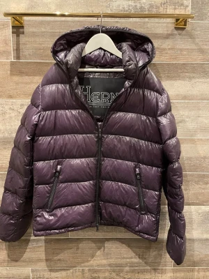 Herno jacka  - Snygg lila dunjacka från Herno med glansig finish och horisontella sömmar. Jackan har två dragkedjeförsedda fickor framtill, huva och dragkedja hela vägen upp. Perfekt för kalla vinterdagar och riktigt lätt att bära.
