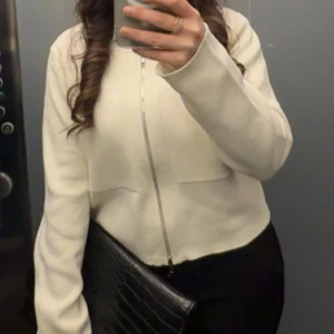 Vit kofta med dragkedja - Snygg vit cardigan med rak passform och lång ärm. Tröjan har en stilren design med dragkedja framtill och är perfekt att matcha med svarta jeans eller kjol. Materialet känns mjukt och bekvämt mot huden, och den neutrala färgen gör den enkel att styla. 