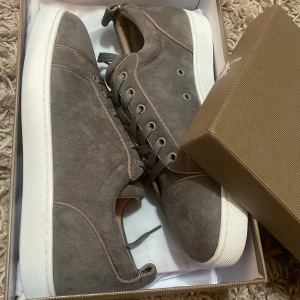 Grå mockasneakers från Louboutin - Snygga grå sneakers i mocka från Louboutin med klassisk röd sula och vita kanter. Skorna har rund tå, snörning och diskreta detaljer i beige. Perfekta för dig som vill ha något stilrent men ändå sticker ut med den ikoniska röda sulan.